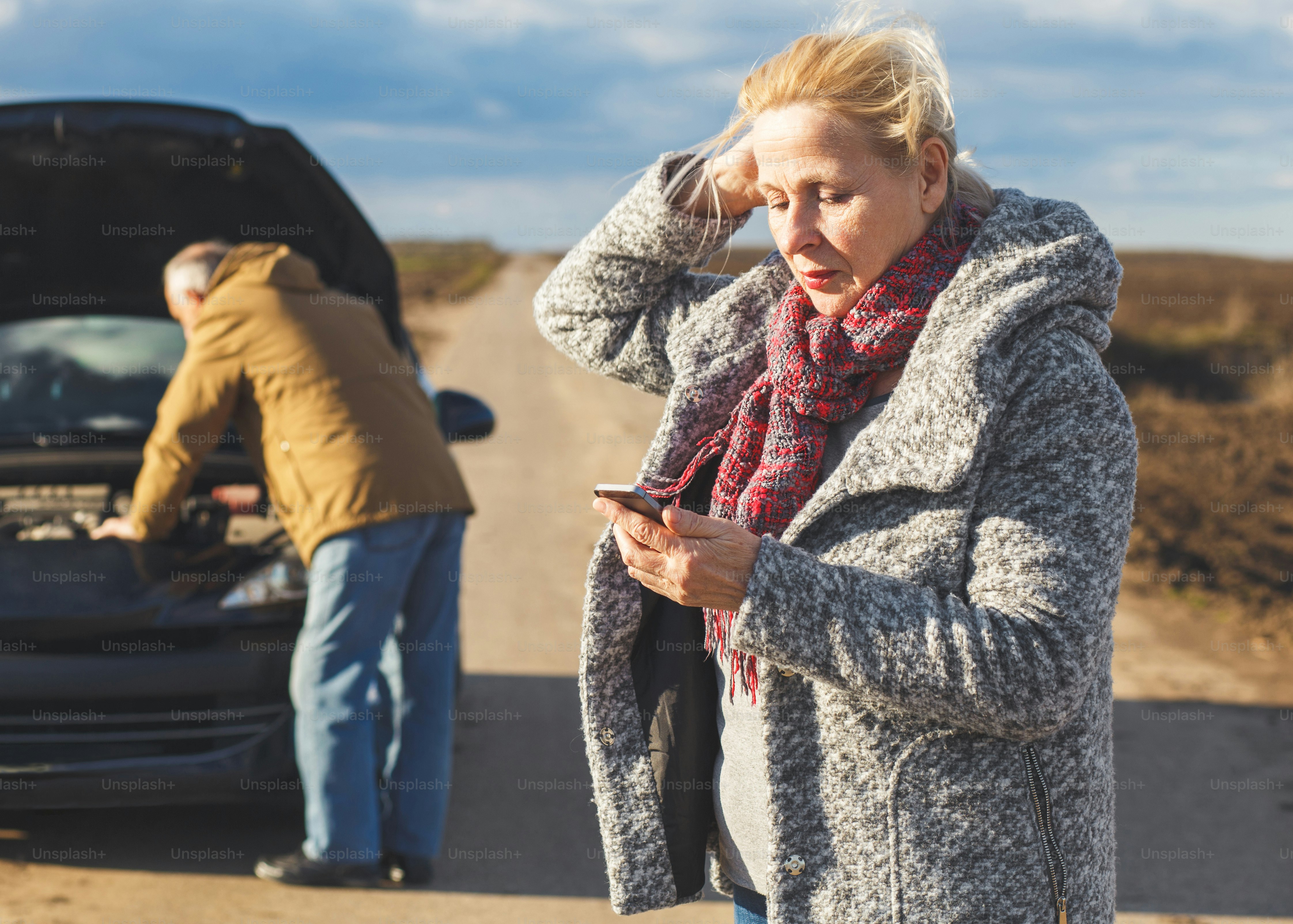 Senior couple on the road having problem with a car, woman calling assistance while man looking at vehicle engine